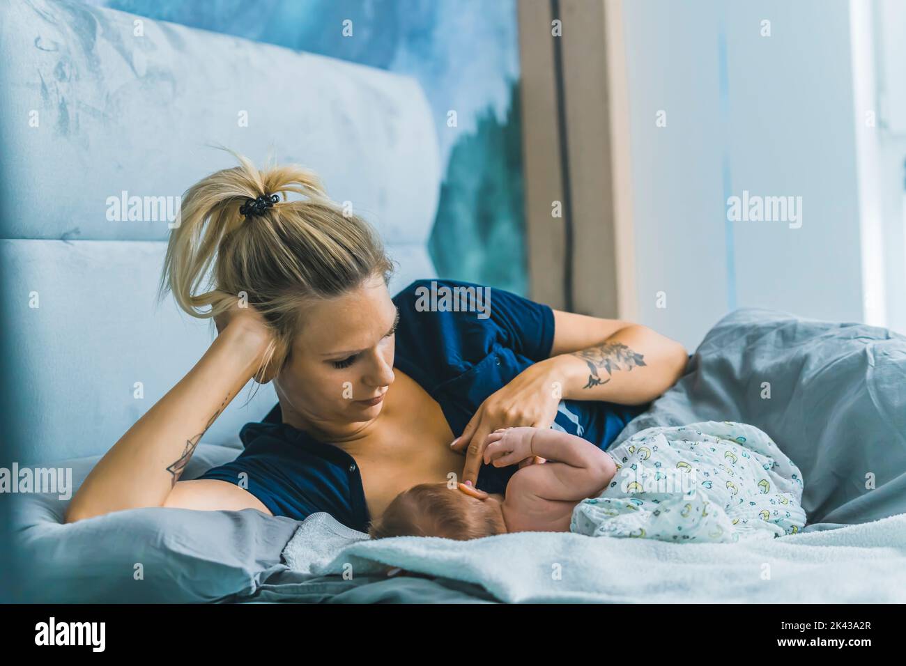 Caucasian young mom laying in bed with her newborn son under a blanket and breastfeeding him in a blue baby boy room. High quality photo Stock Photo
