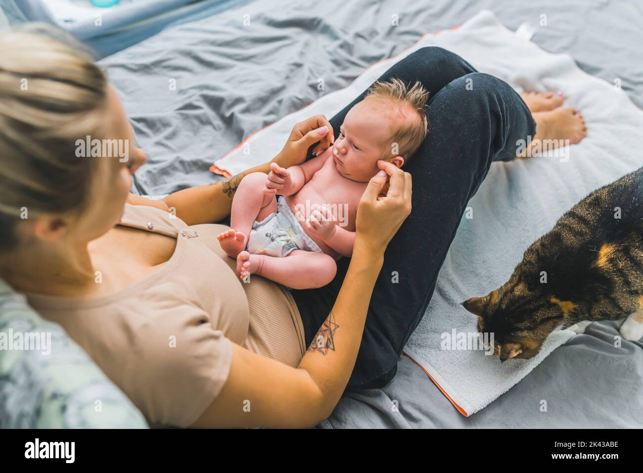 Young mother sitting on a bed, holding her newborn son with cute brown hair on her lap, communicating with him. Big multicolored cat sitting near them and sniffing the area. High quality photo Stock Photo