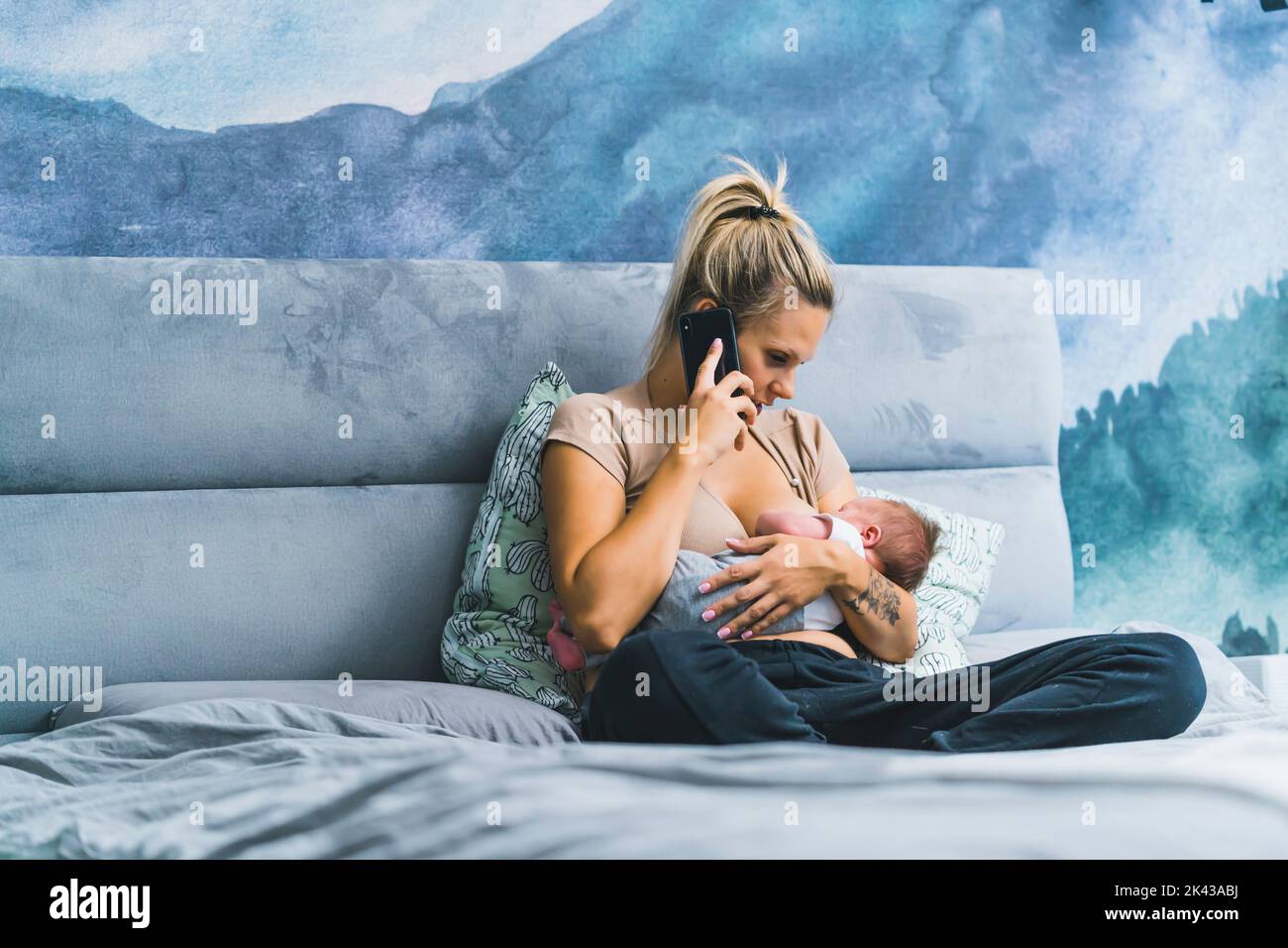 Happy young successful mother sitting on a bed and breastfeeding her infant son while having a work phone call. High quality photo Stock Photo