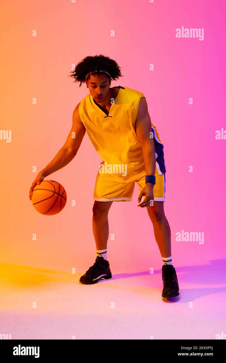 Image of biracial basketball player bouncing basketball on pink to orange background. Sports and competition concept. Stock Photo