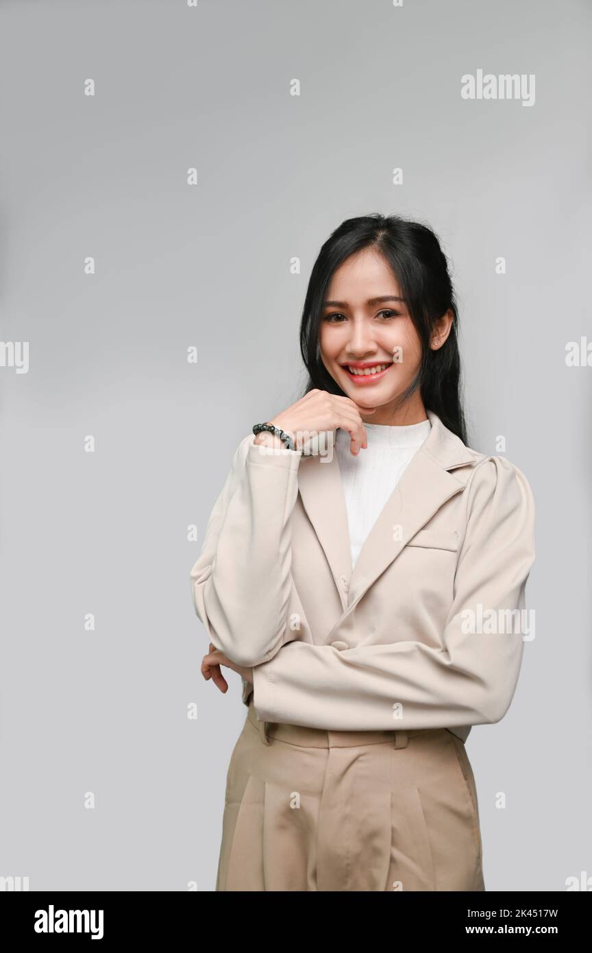 Portrait, Attractive gorgeous young Asian female or businesswoman in casual suit smiling, hand on chin, standing isolated over grey studio background. Stock Photo