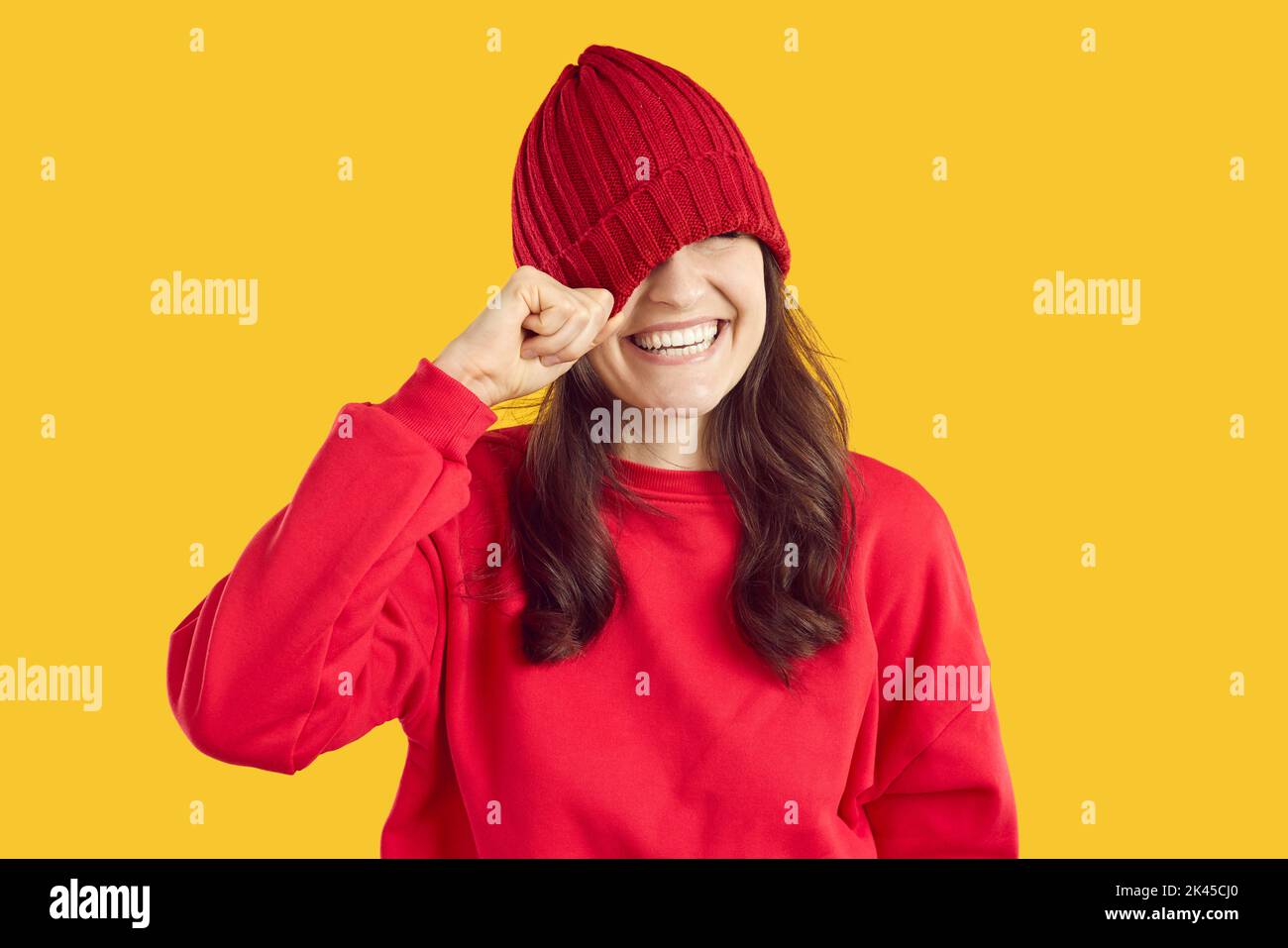 Funny cute young woman laughs and hides her eyes by pulling knitted hat over them. Stock Photo