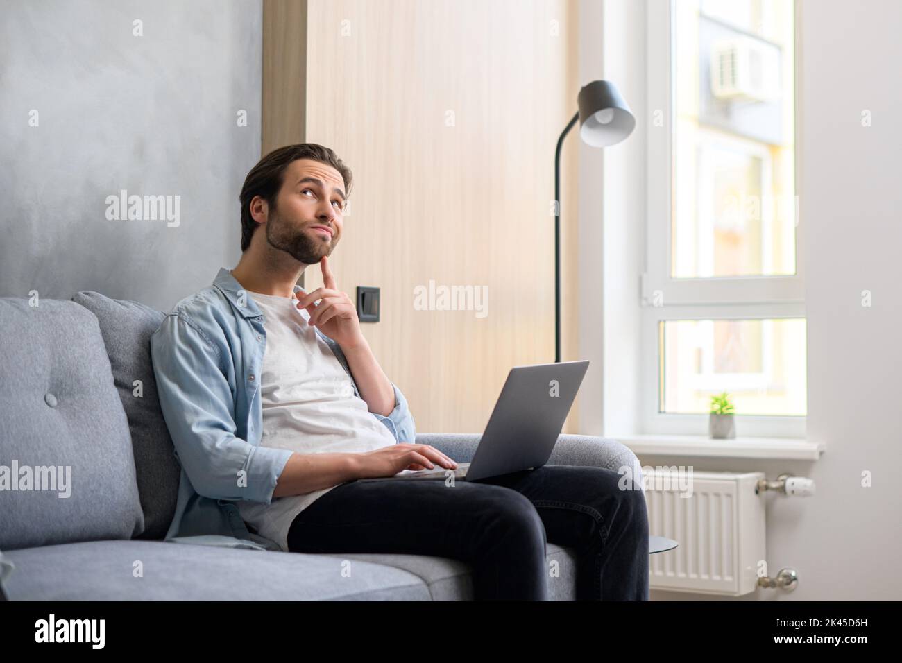 Pensive young businessman in casual wear sitting at the sofa at home, using laptop computer, watch away thoughtfully, looking for business tasks solving. Male employee on the couch Stock Photo