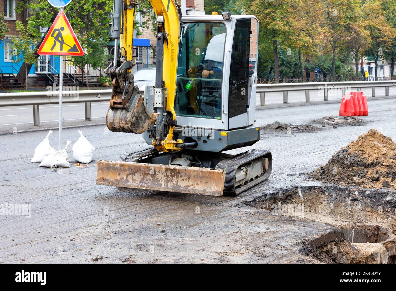 A small road excavator at work repairing the city's sewer infrastructure on a fenced roadway. Copy space. Stock Photo