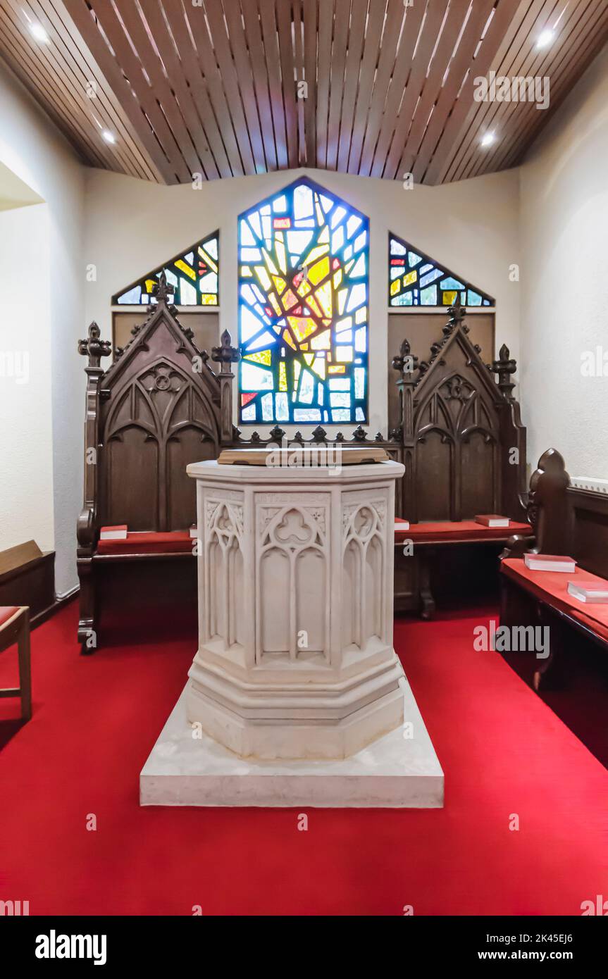 Baptismal font and two wooden chairs, with a modern stained glass window inside a church. Stock Photo