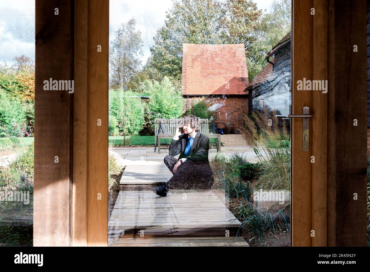 Man sitting on path to house, seen through glass door. Stock Photo