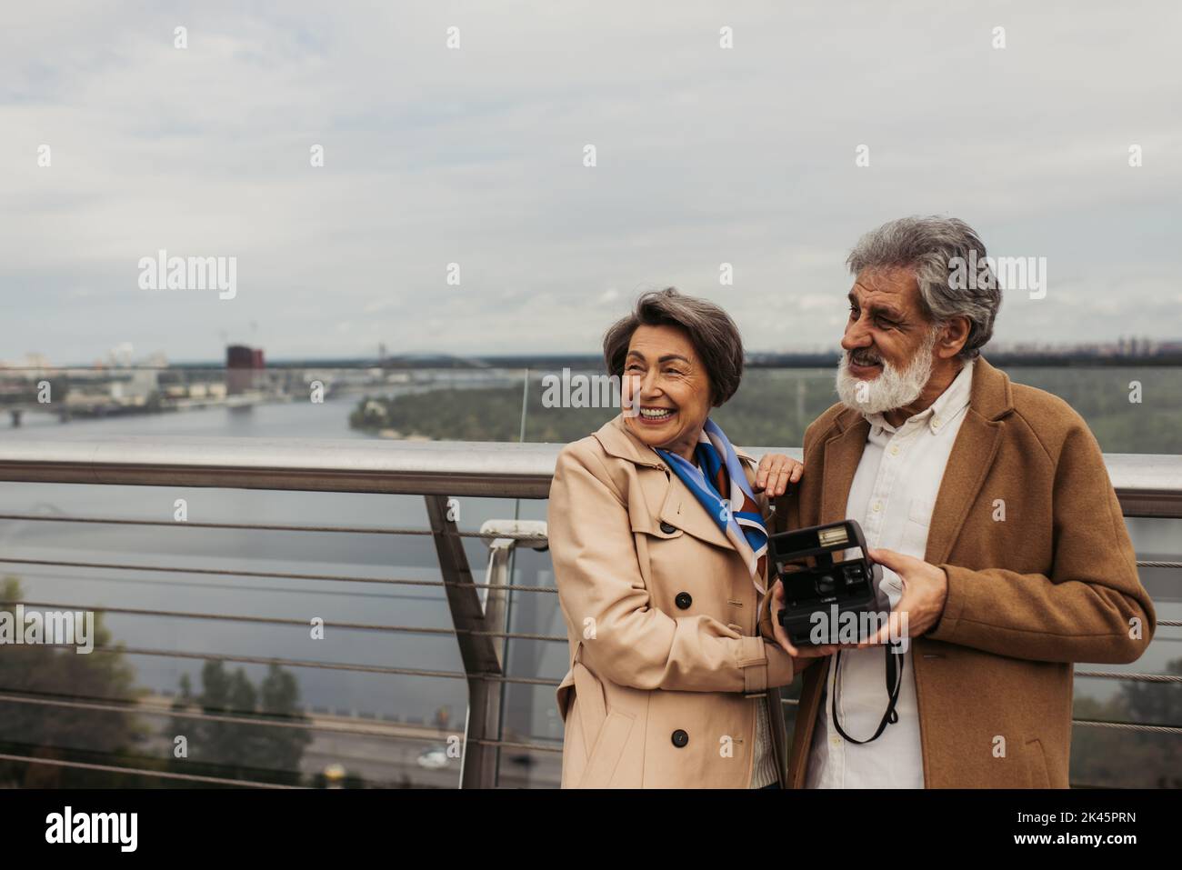 happy senior woman in trench coat hugging husband with vintage camera ...