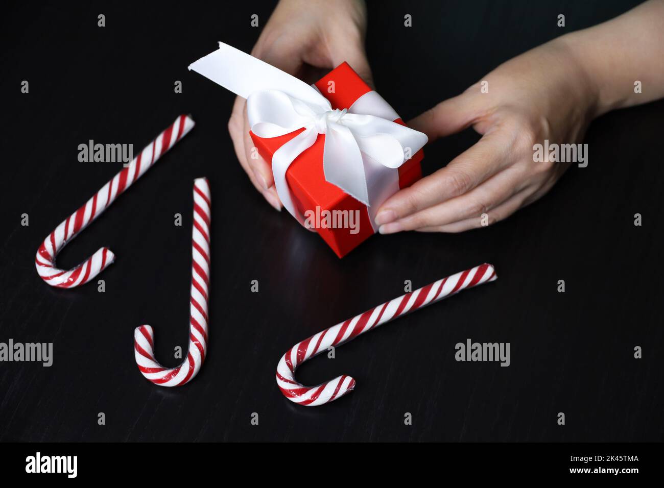 Red gift box in female hands on dark wooden table with candy canes. Concept of Christmas holiday, romantic surprise Stock Photo