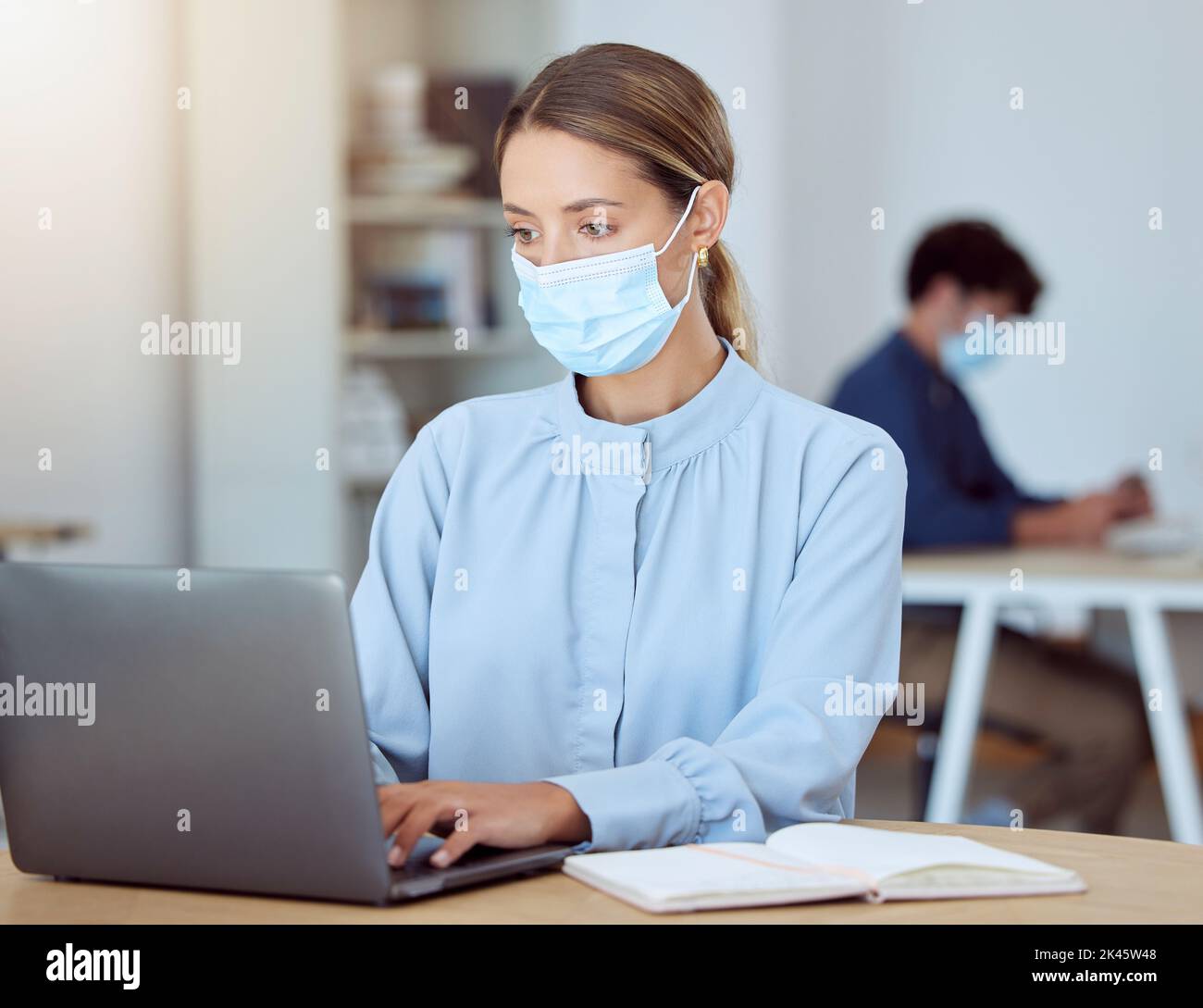 Laptop, face mask and corporate employee working on a project while sitting at a desk in the office. Professional woman typing company documents on Stock Photo