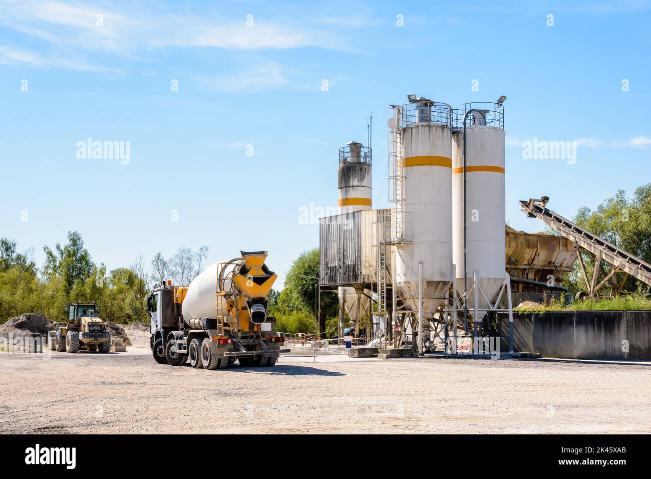 A concrete mixer truck and a wheel loader are parked next to sand silos in a quarry on a sunny day. Stock Photo