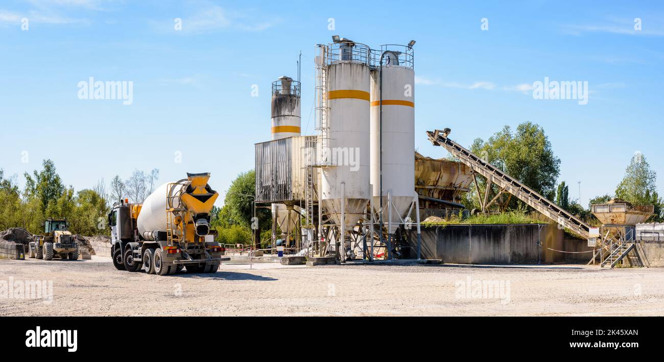 A concrete mixer truck and a wheel loader are parked next to sand silos in a quarry on a sunny day. Stock Photo
