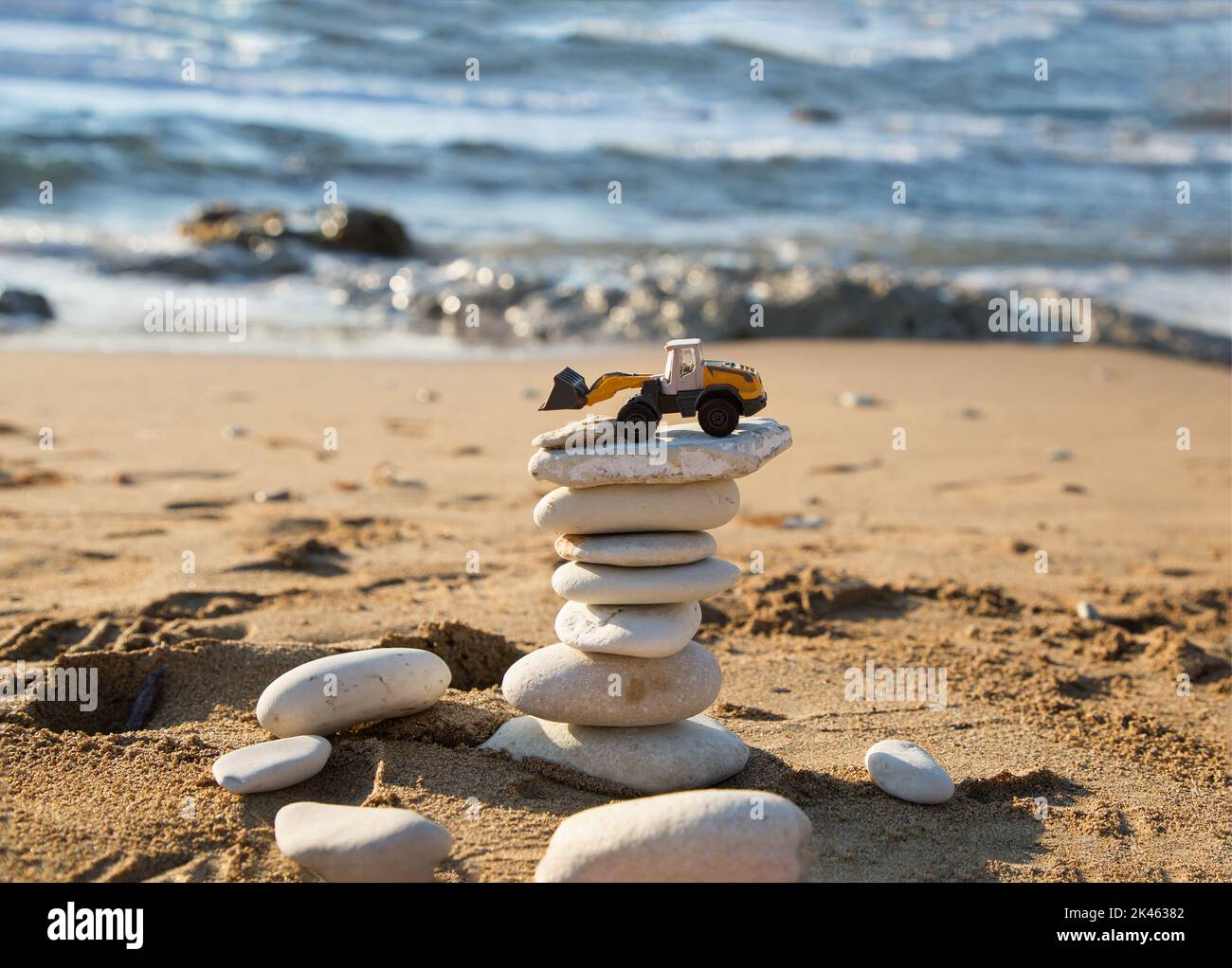 orange mini toy backhoe loader stands on tower of flat stones against backdrop of the sea. Vacation in a construction company. The concept of stabilit Stock Photo