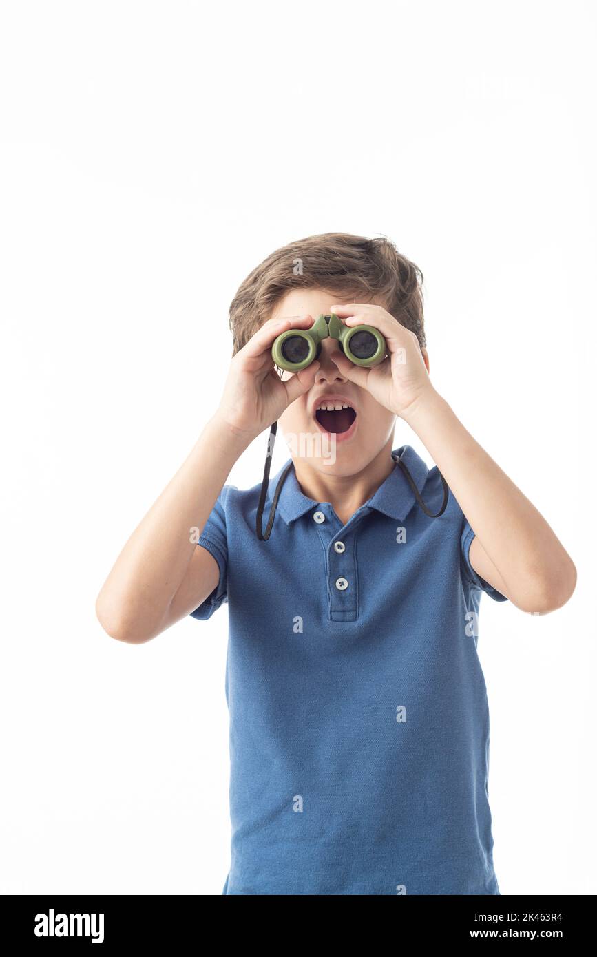 Caucasian boy in blue T-shirt looking at camera with surprised expression through binoculars, on white background. Stock Photo