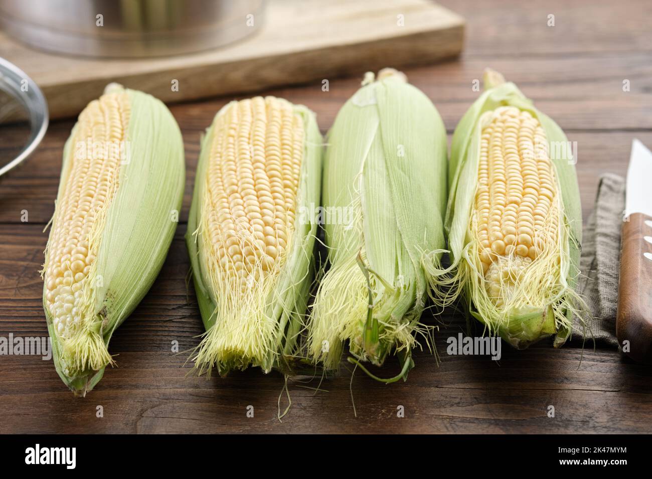 Raw sweet corn cobs for cooking and kitchen knife on kitchen table, cooking pot on background. Stock Photo