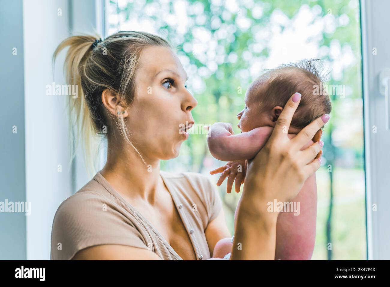 Caucasian happy loving mother holding her first child, tiny newborn son, in the air and playing with him, showing grice and emotions while standing near the window at home. High quality photo Stock Photo