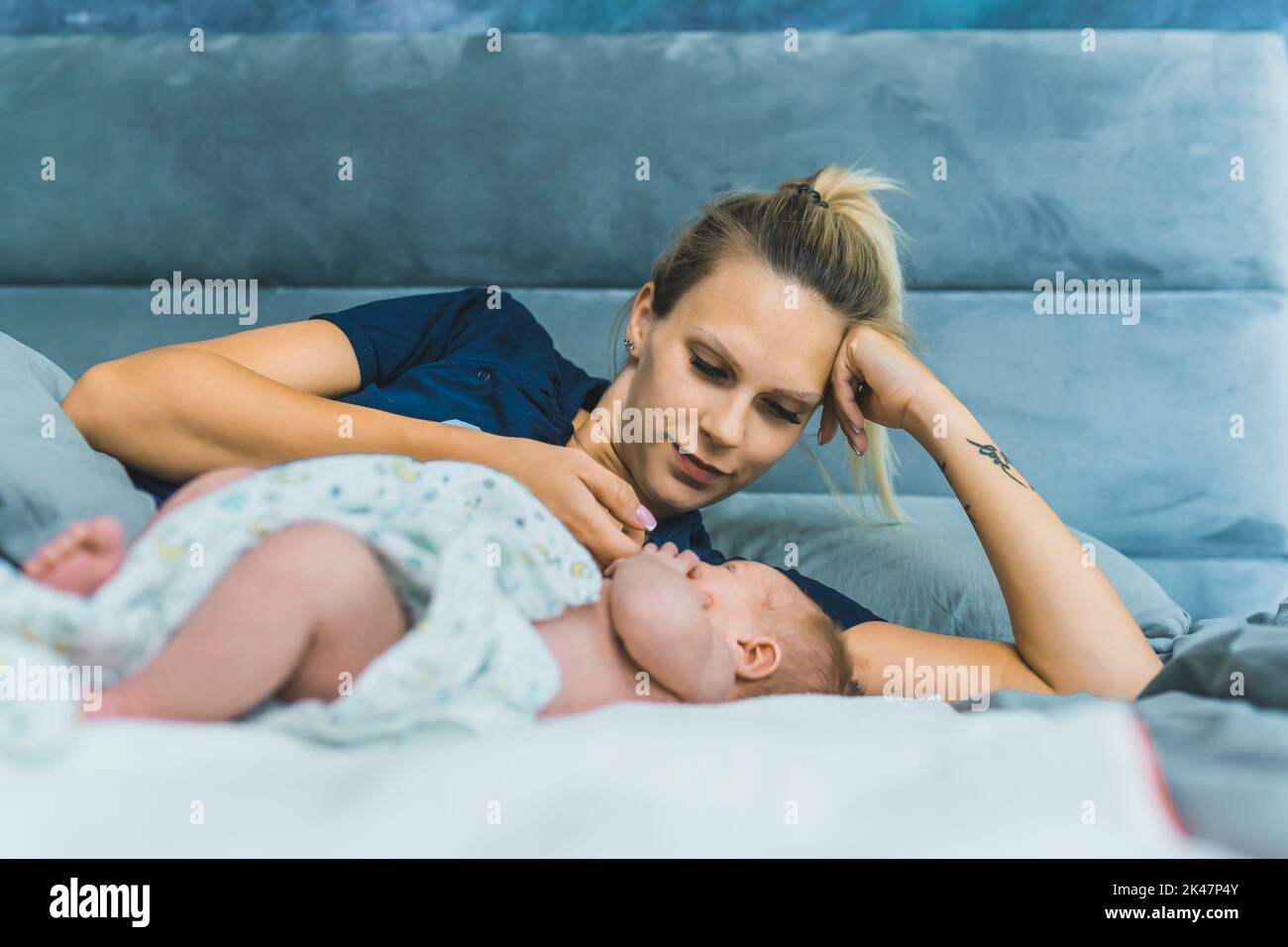 Caucasian beautiful young mom laying in bed with her newborn son wrapped in a baby blanket, talking to him, enjoying precious time with her child. High quality photo Stock Photo