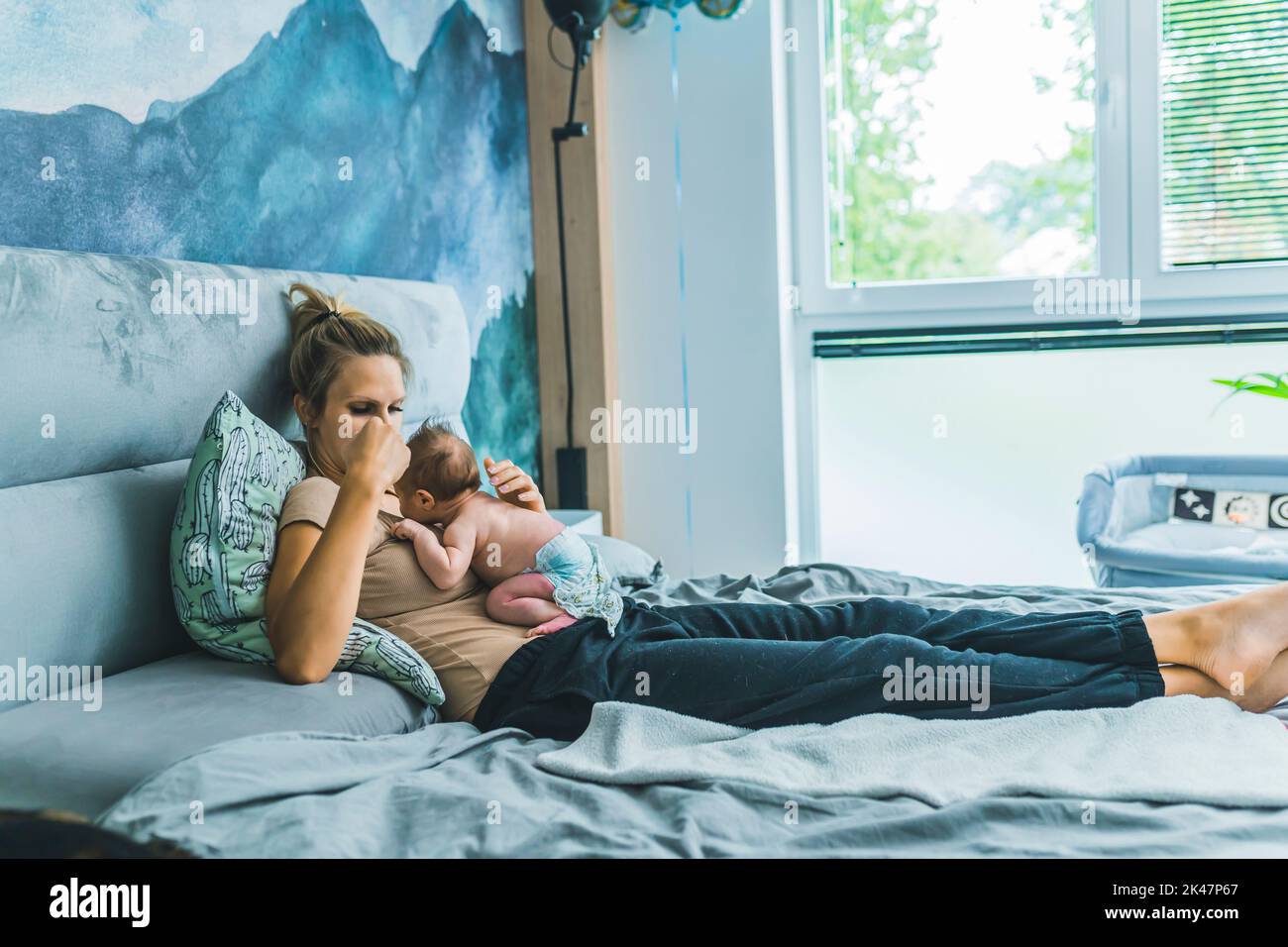 Caucasian beautiful young mom relaxing in bed and her newborn son in a diaper laying in a fetal position on her stomach in a designed nursery room in blue colors. High quality photo Stock Photo