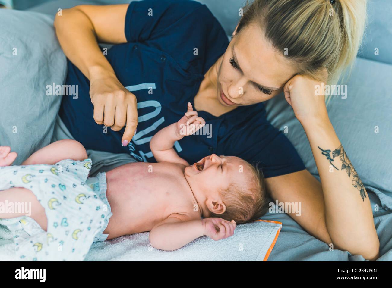 Caucasian young mom laying in bed with her couple months old baby boy. Her son is wearing a diaper, screaming and crying and she is trying to calm him down. High quality photo Stock Photo
