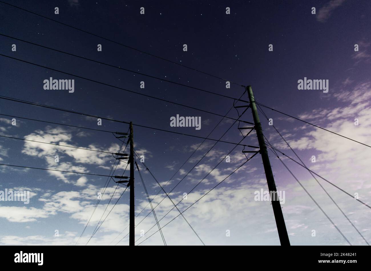 two power poles with power lines seen throughout taken at night with a few stars visible in the background behind the poles. Stock Photo