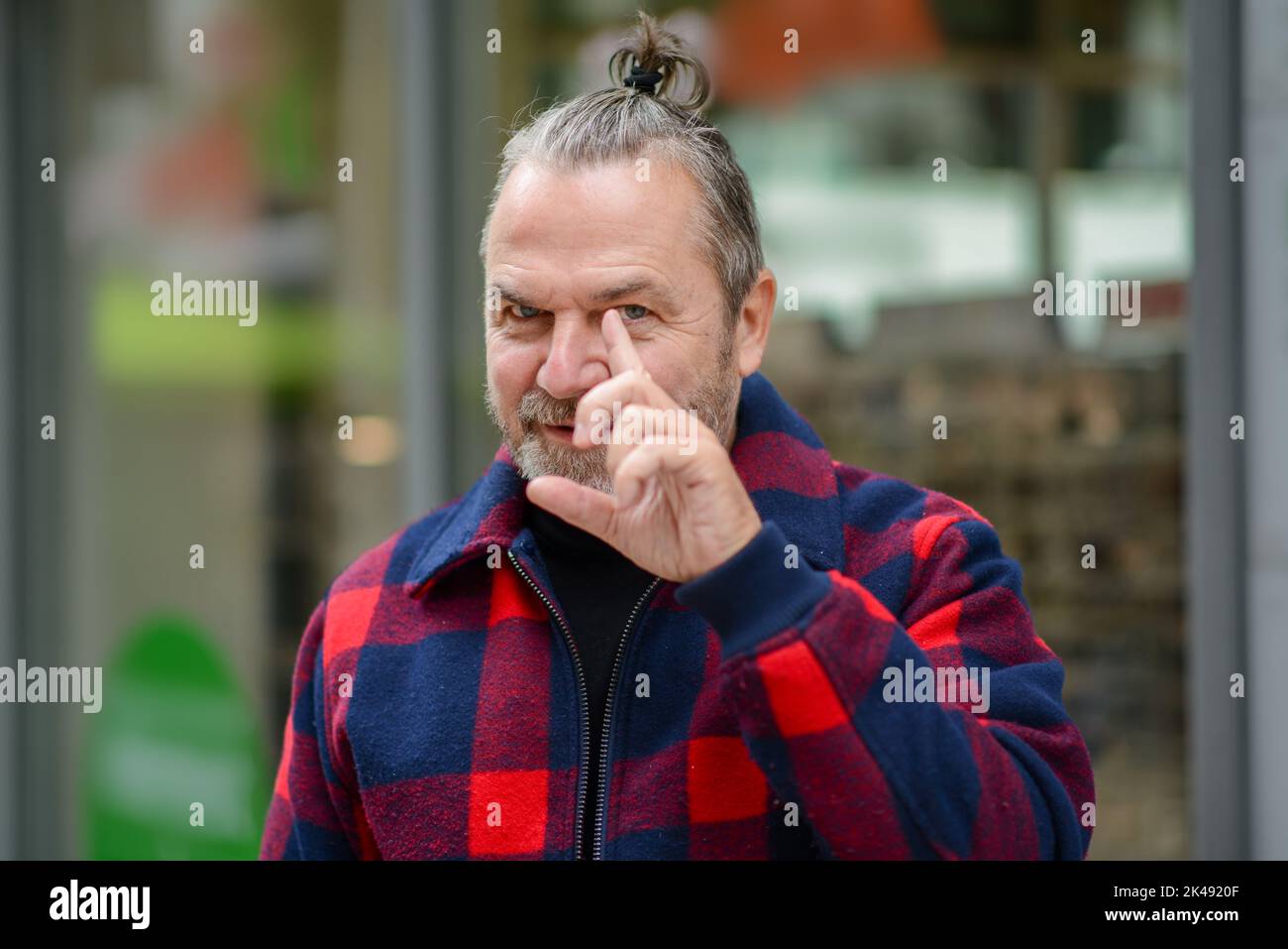 Middle aged man with a messy bun in a red and blue lumberjack style jacket is standing in a shopping street and pointing to his front eye Stock Photo