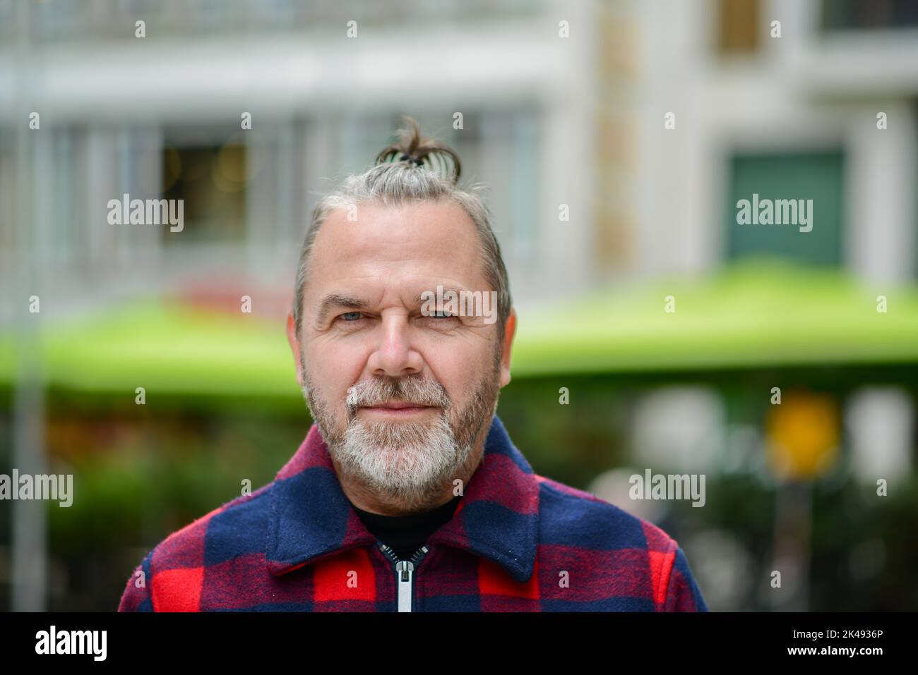 Middle aged man with a messy bun in a red and blue lumberjack style jacket is standing in a shopping street with a soft smile Stock Photo