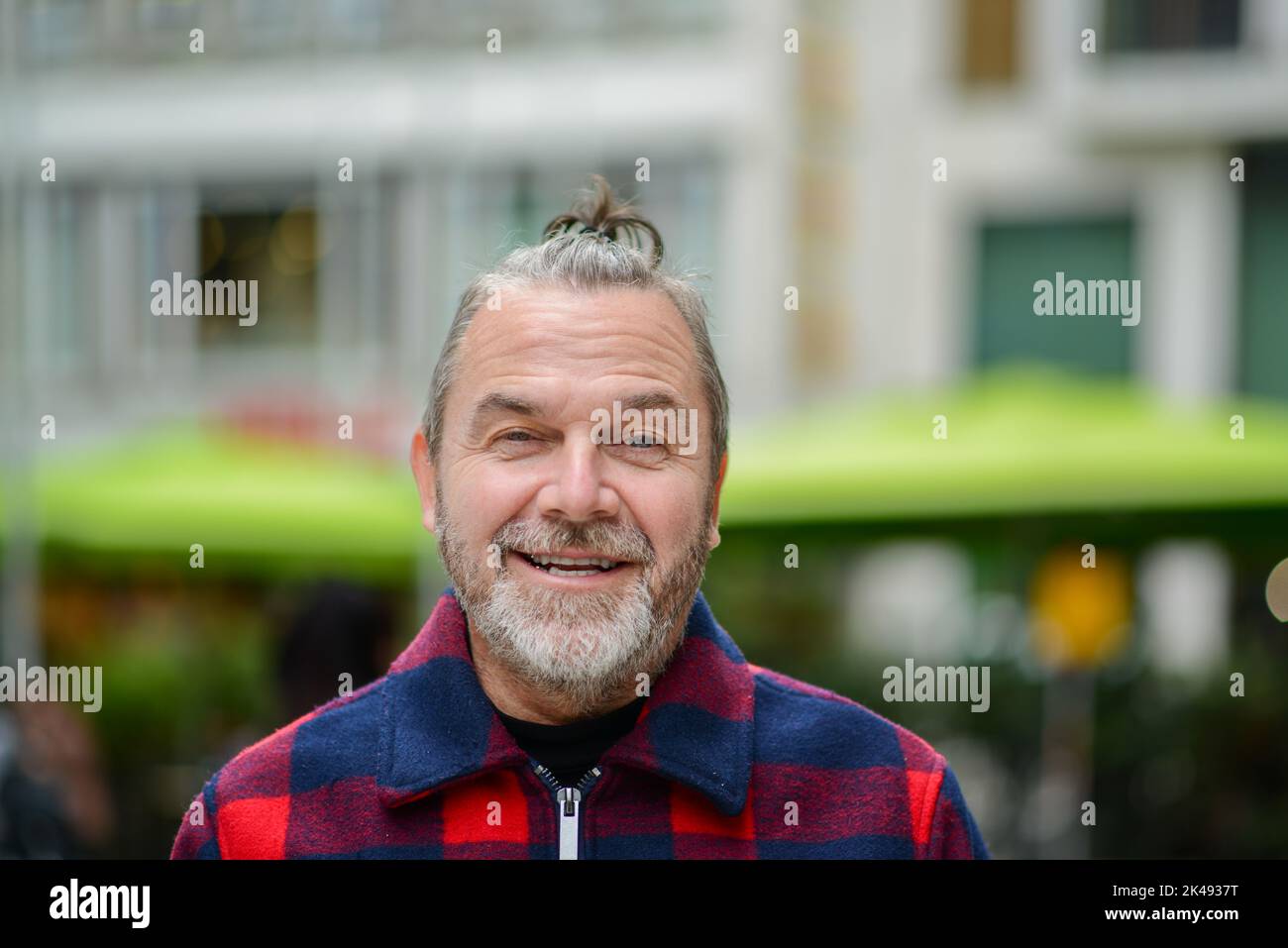 Middle aged man with a messy bun in a red and blue lumberjack style jacket is standing in a shopping street with a mischievous smile Stock Photo