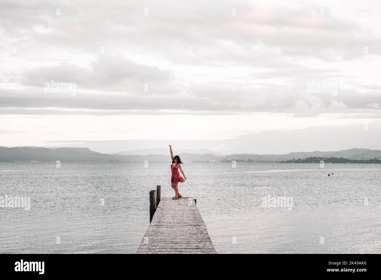 Young caucasian girl wearing sandals glasses and a pink dress dancing with her arm raised on the lake pier under the clouds in Okere, new zealand Stock Photo