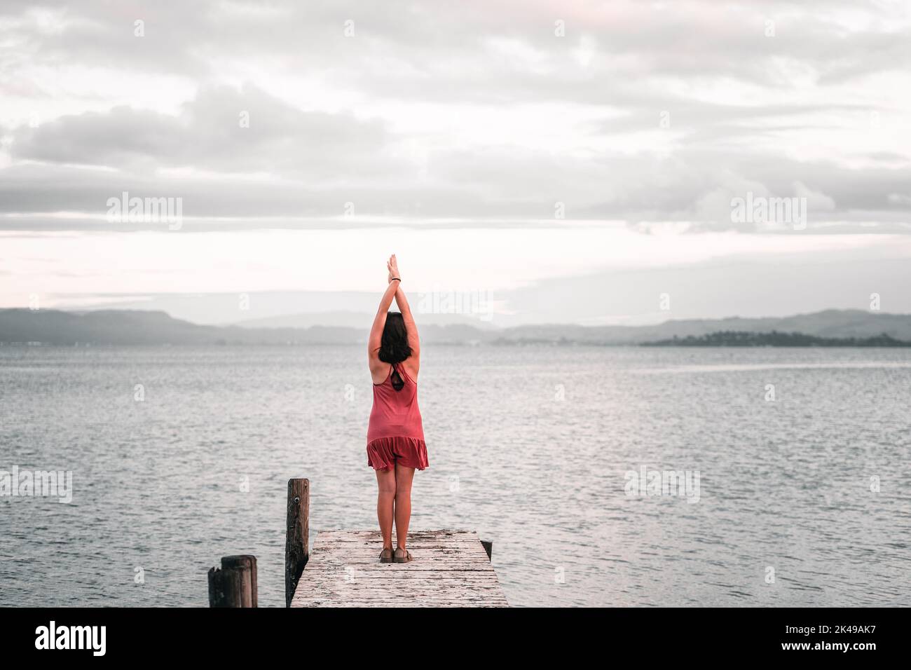 Caucasian young woman wearing black bracelet pink dress and sandals standing with feet together and arms outstretched upwards at end of wooden pier Stock Photo