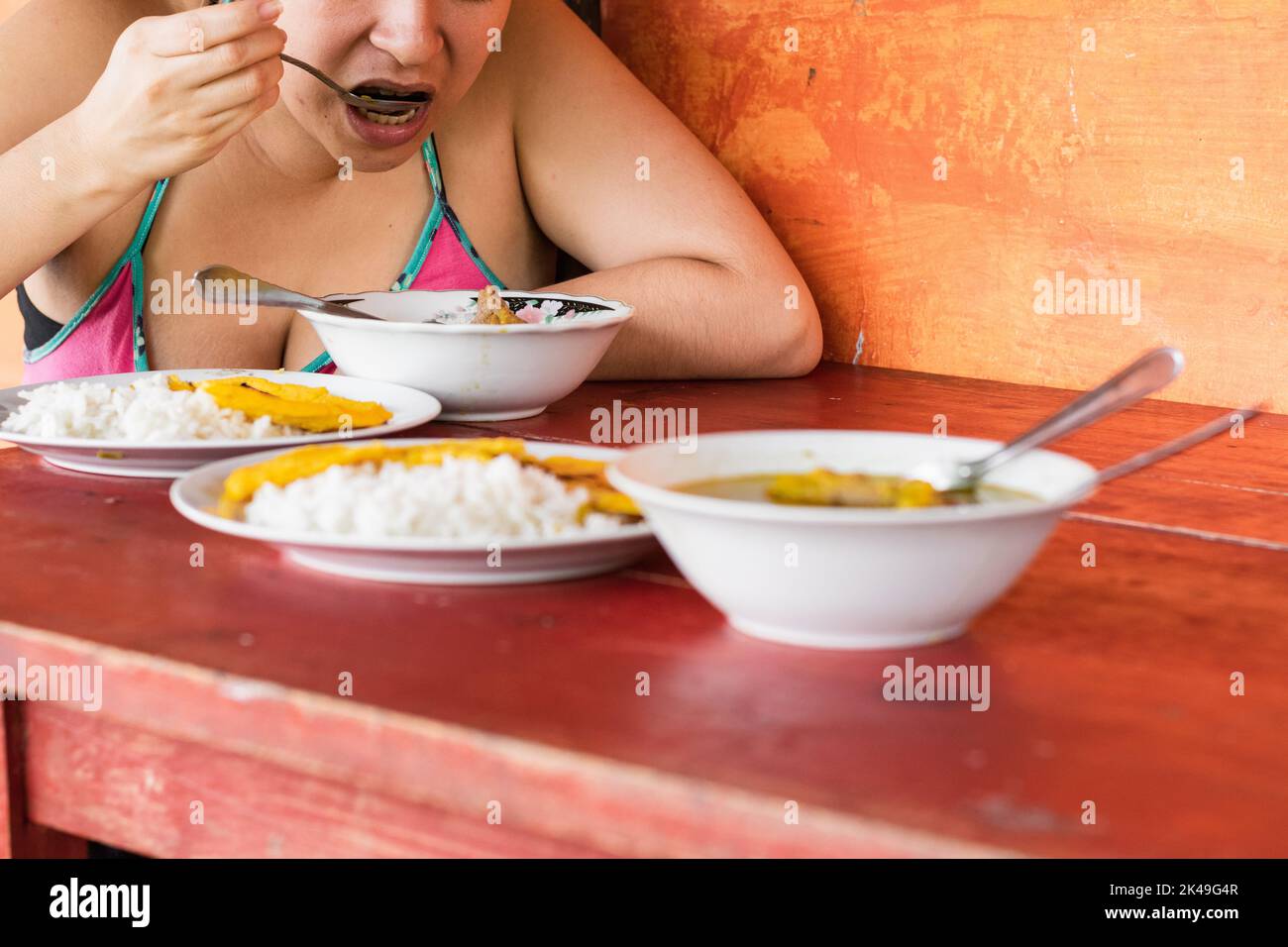 close-up of a latin peasant woman, eating a typical colombian dish for breakfast. girl holding spoon to mouth, drinking beef broth with yellow arepa a Stock Photo