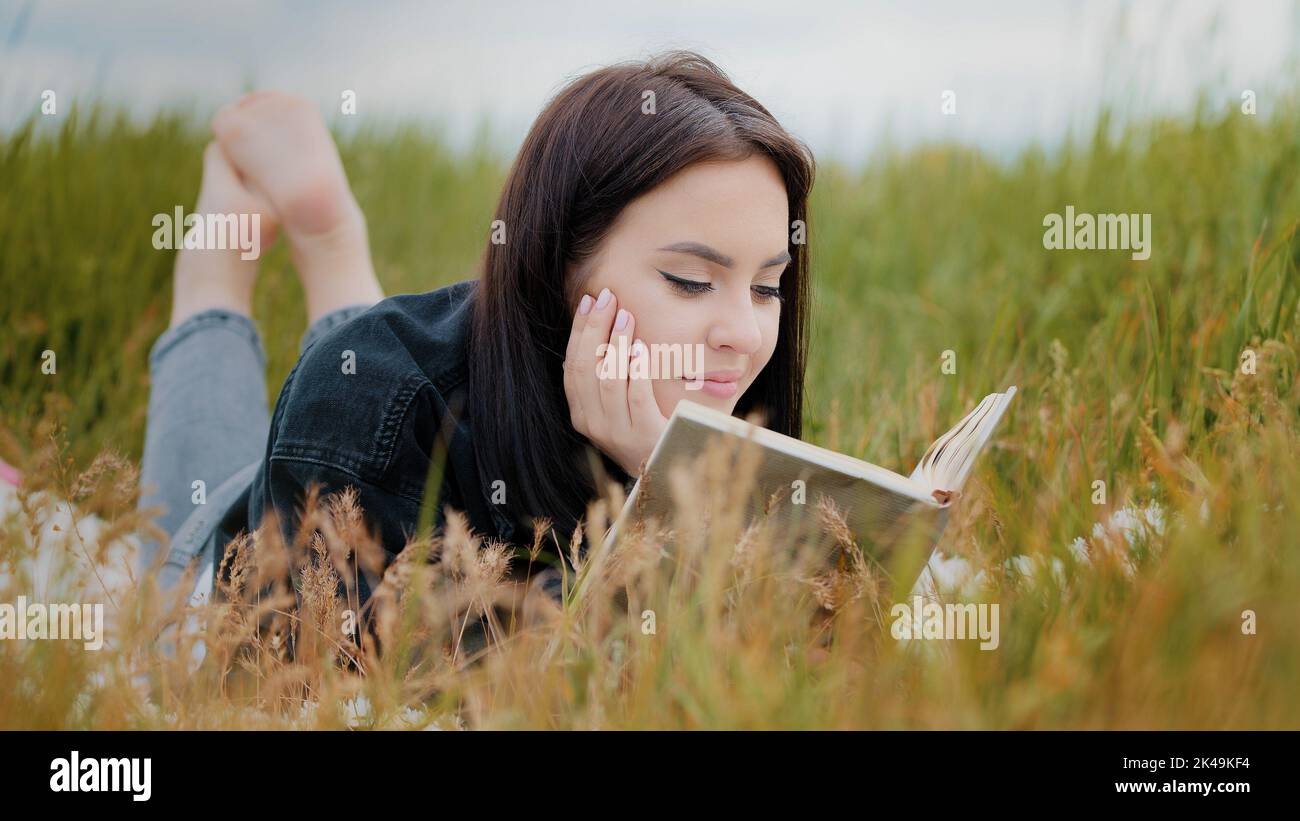 Young attractive brunette caucasian woman girl lying on grass pretty teenage girlfriend student read interesting book educational literature novel Stock Photo
