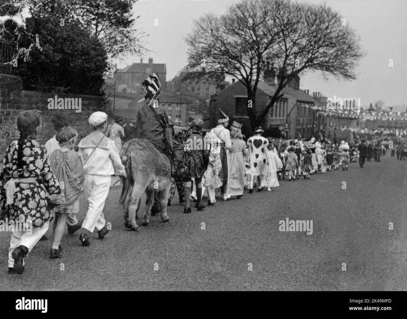Children in fancy dress, including a boy riding a donkey, walk in procession on 6 May 1935, the Silver Jubilee Day of King George V, down a long straight street (probably Wakefield Road) decked with flags and bunting at Denby Dale in West Yorkshire, England, UK. Stock Photo