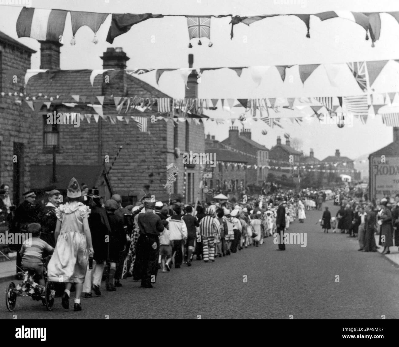 Schoolchildren wearing fancy dress costume walk in procession on 6 May 1935, the Silver Jubilee Day of King George V, down a long straight street (probably Wakefield Road) decked with Union flags and bunting at Denby Dale in West Yorkshire, England, UK. Stock Photo