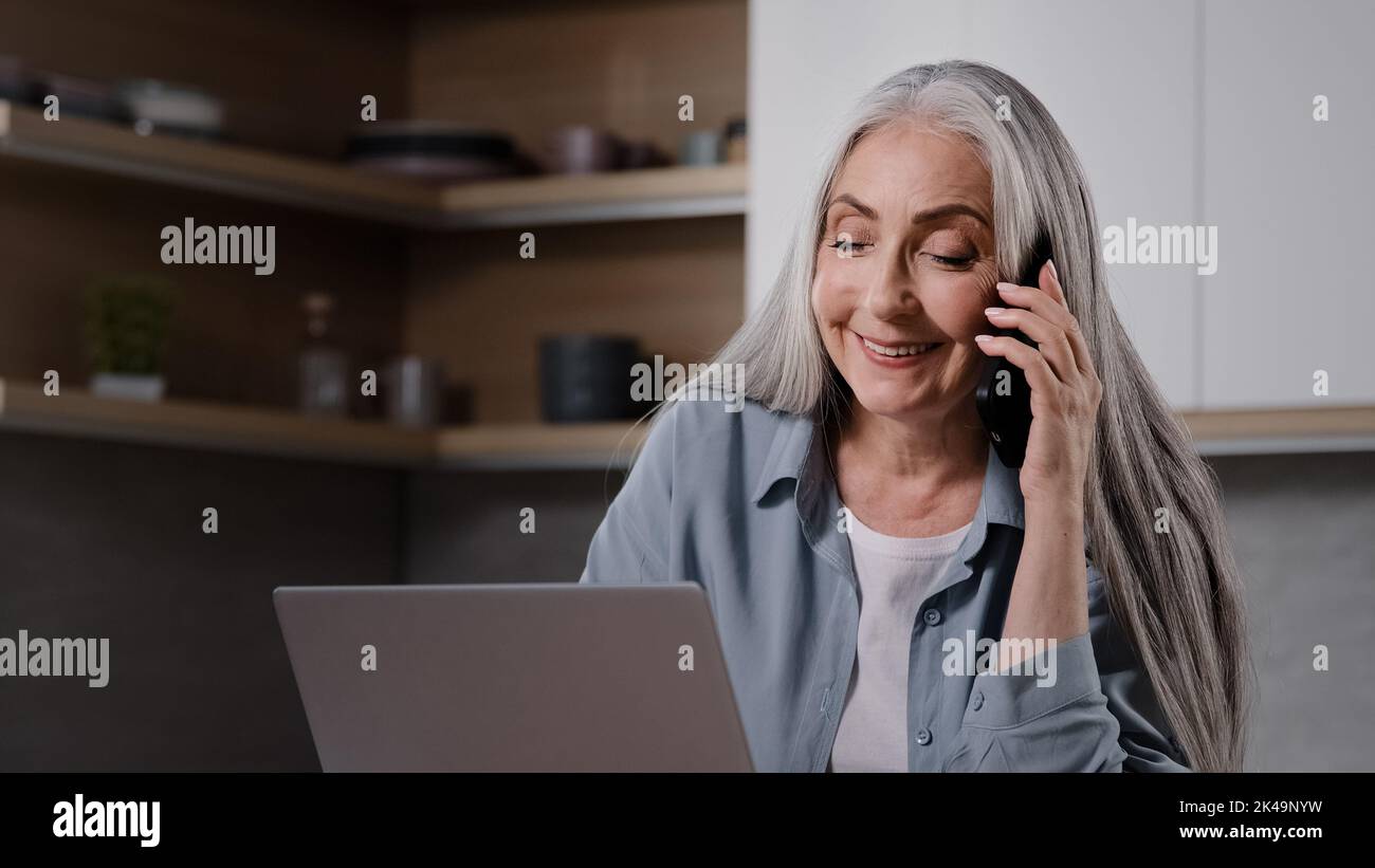 Female senior businesswoman middle aged grey-haired woman specialist freelancer sit in kitchen negotiating with business partner remotely by phone Stock Photo