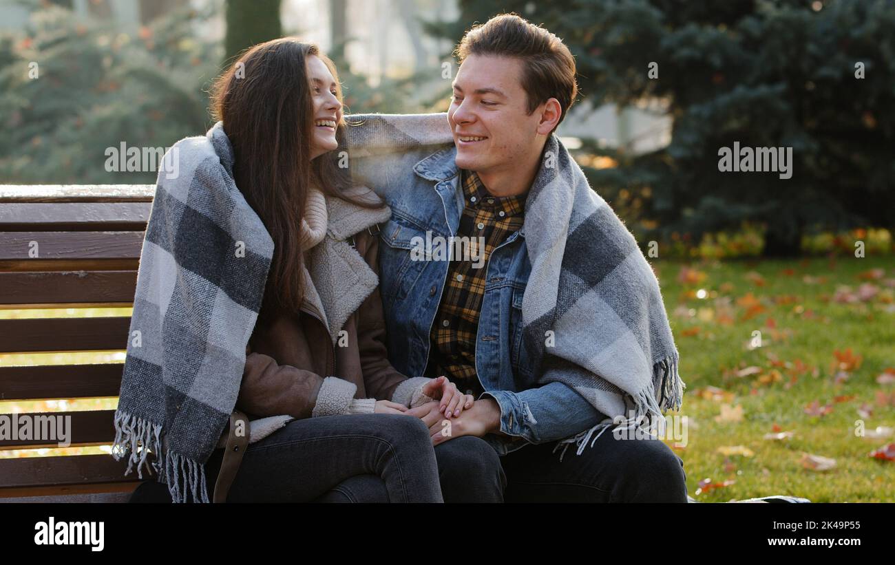 Cute loving couple sit on bench warming each other covering wool blanket outdoors in cold froze autumnal weather feel care love hugging funny talking Stock Photo