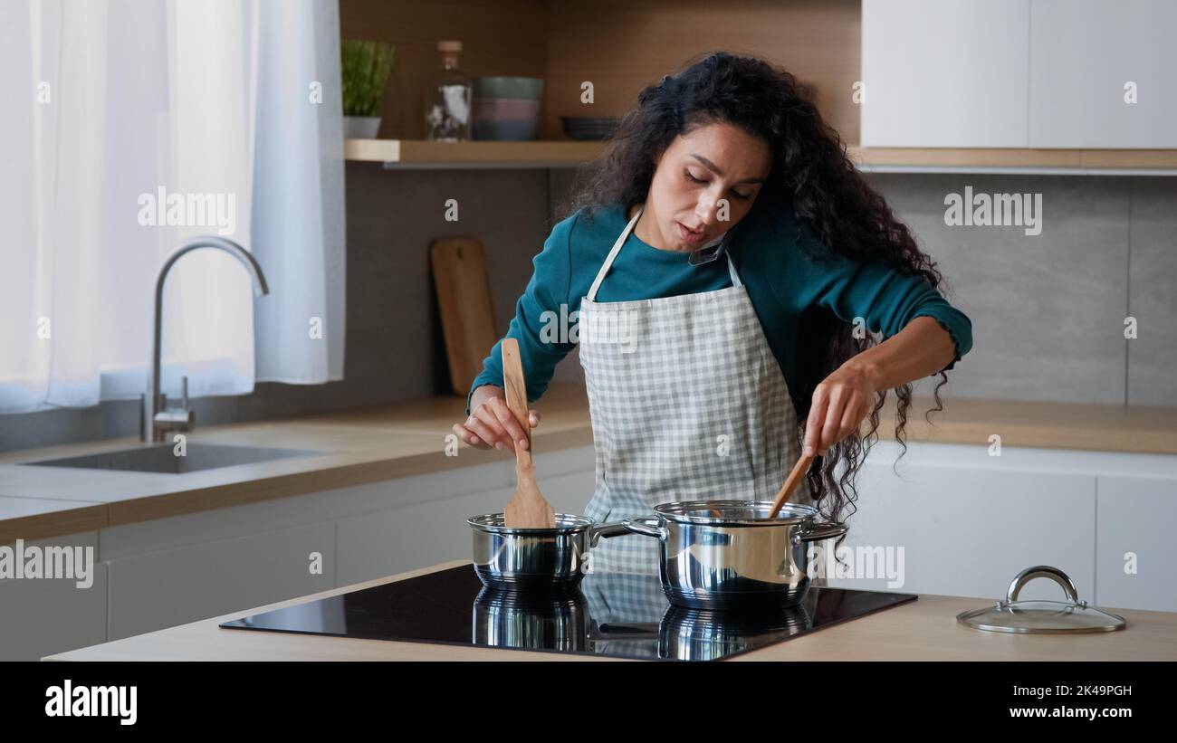 Busy arabian mom attractive curly housewife chef wears apron cooking breakfast at home kitchen stirring meal in saucepans with spoon preparing food Stock Photo