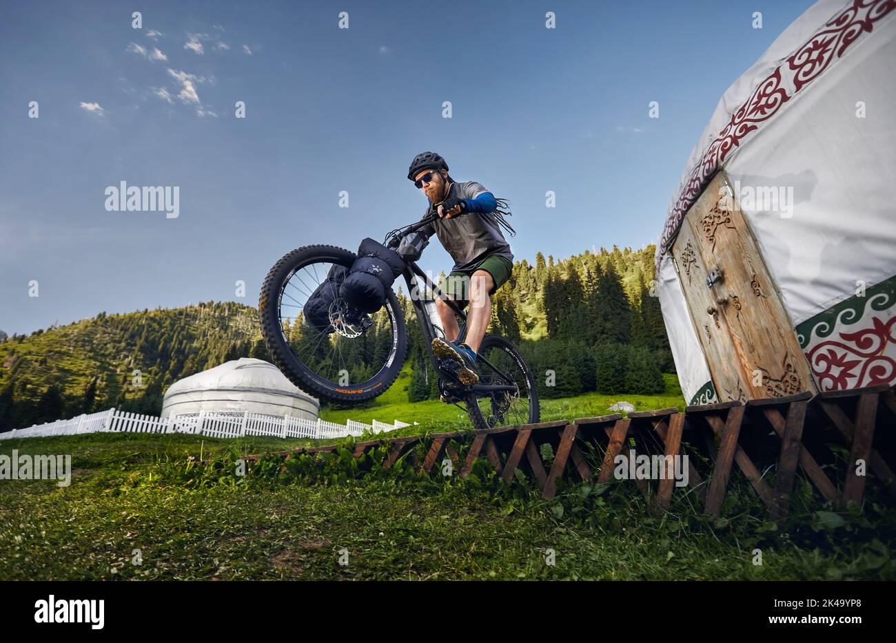 Man with dreadlocks and beard ride and jump at mountain bike with tourist bags near Nomad Yurt house in beautiful mountain valley in Almaty, Kazakhsta Stock Photo