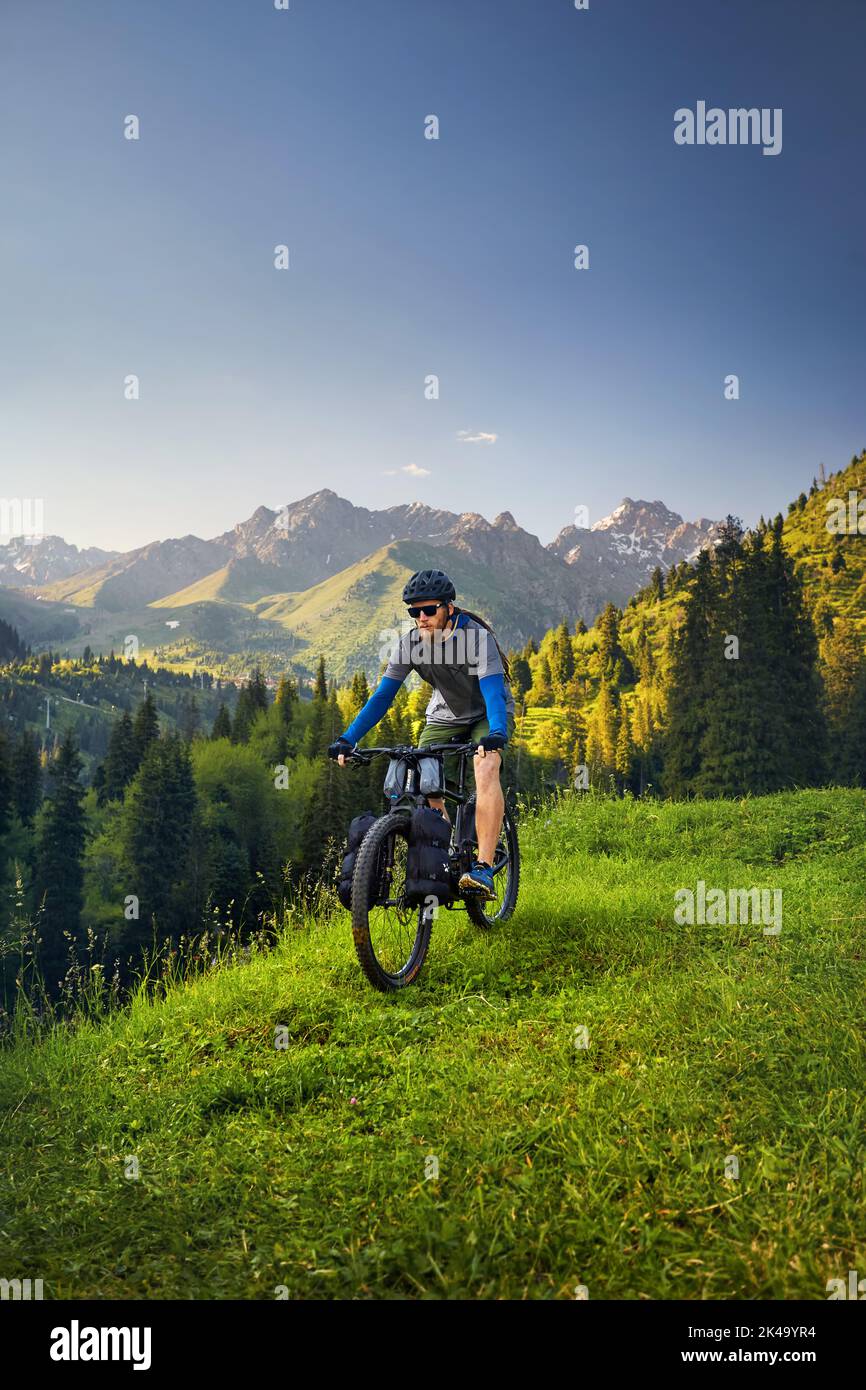 Man with dreadlocks and beard ride mountain bike with tourist bags at the green hill forest in beautiful mountain valley at sunset in Almaty, Kazakhst Stock Photo