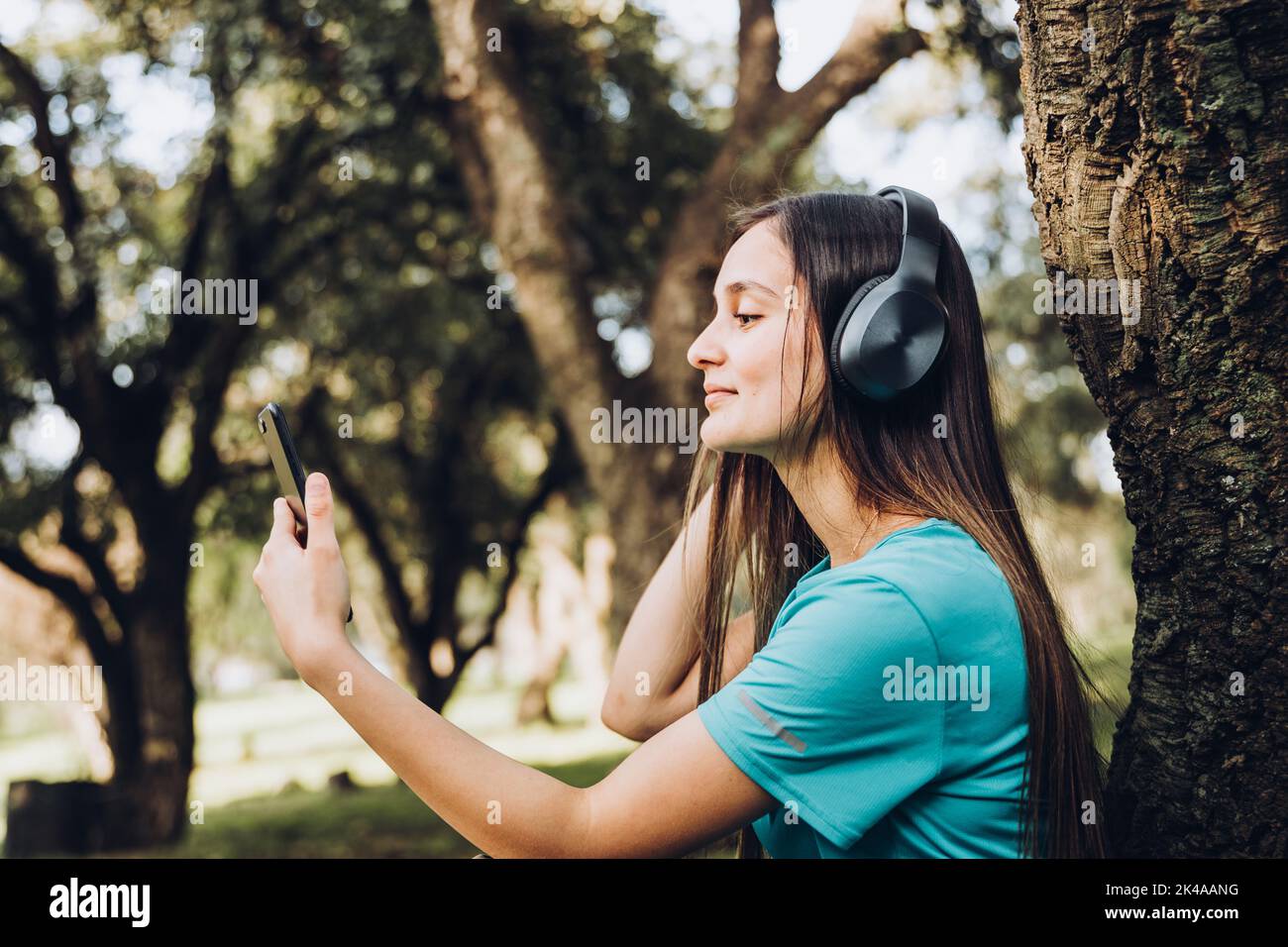 Sitting smiling teenage girl using headphones and listening music on her smartphone in the forest in the afternoon Stock Photo