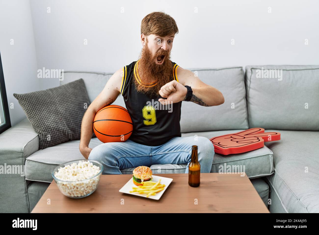 Caucasian man with long beard holding basketball ball cheering tv game looking at the watch time worried, afraid of getting late Stock Photo