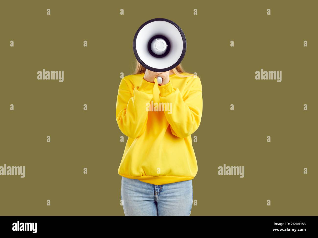 Young woman makes loud announcement hiding her face behind megaphone on khaki background in studio. Stock Photo