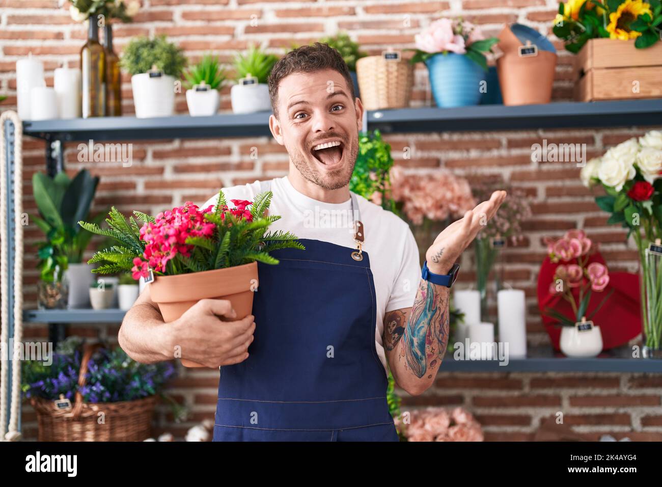Young hispanic man working at florist shop holding plant pot celebrating achievement with happy smile and winner expression with raised hand Stock Photo