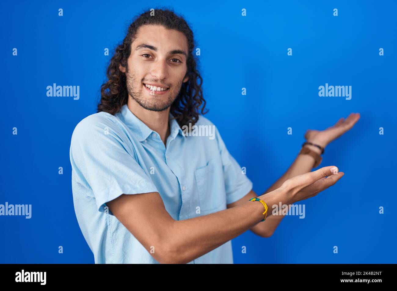 Young hispanic man standing over blue background inviting to enter ...