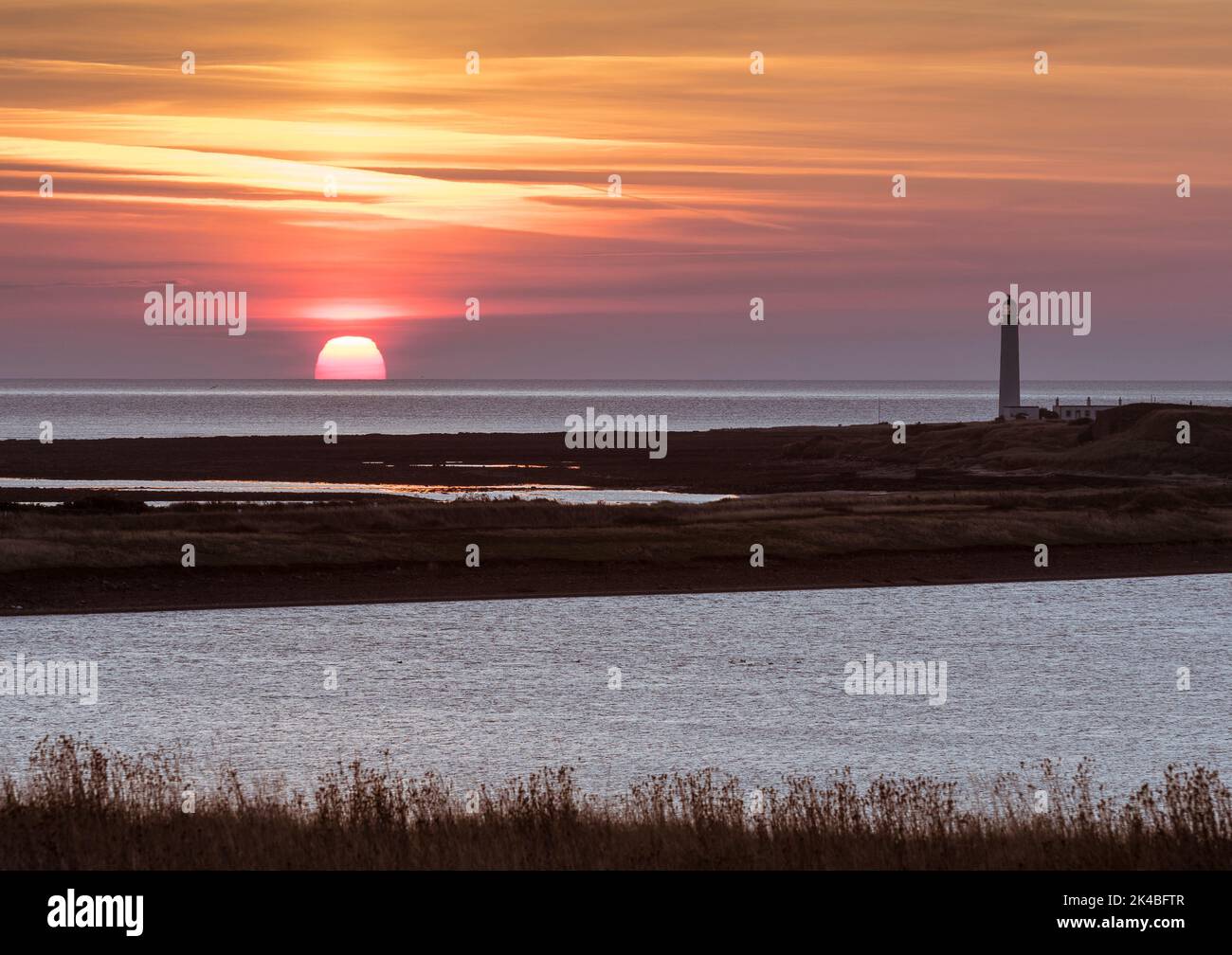 Golden orb of sun partially risen on the sea horizon viewed from Barns Ness point with lighthouse and inshore lagoon in foreground. Stock Photo