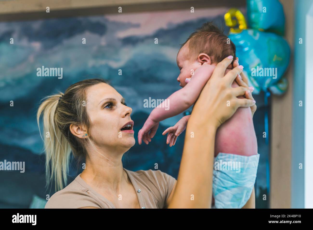 Caucasian happy loving mom holding her tiny newborn son in the air while standing in a blue-designed baby boy room at home. High quality photo Stock Photo