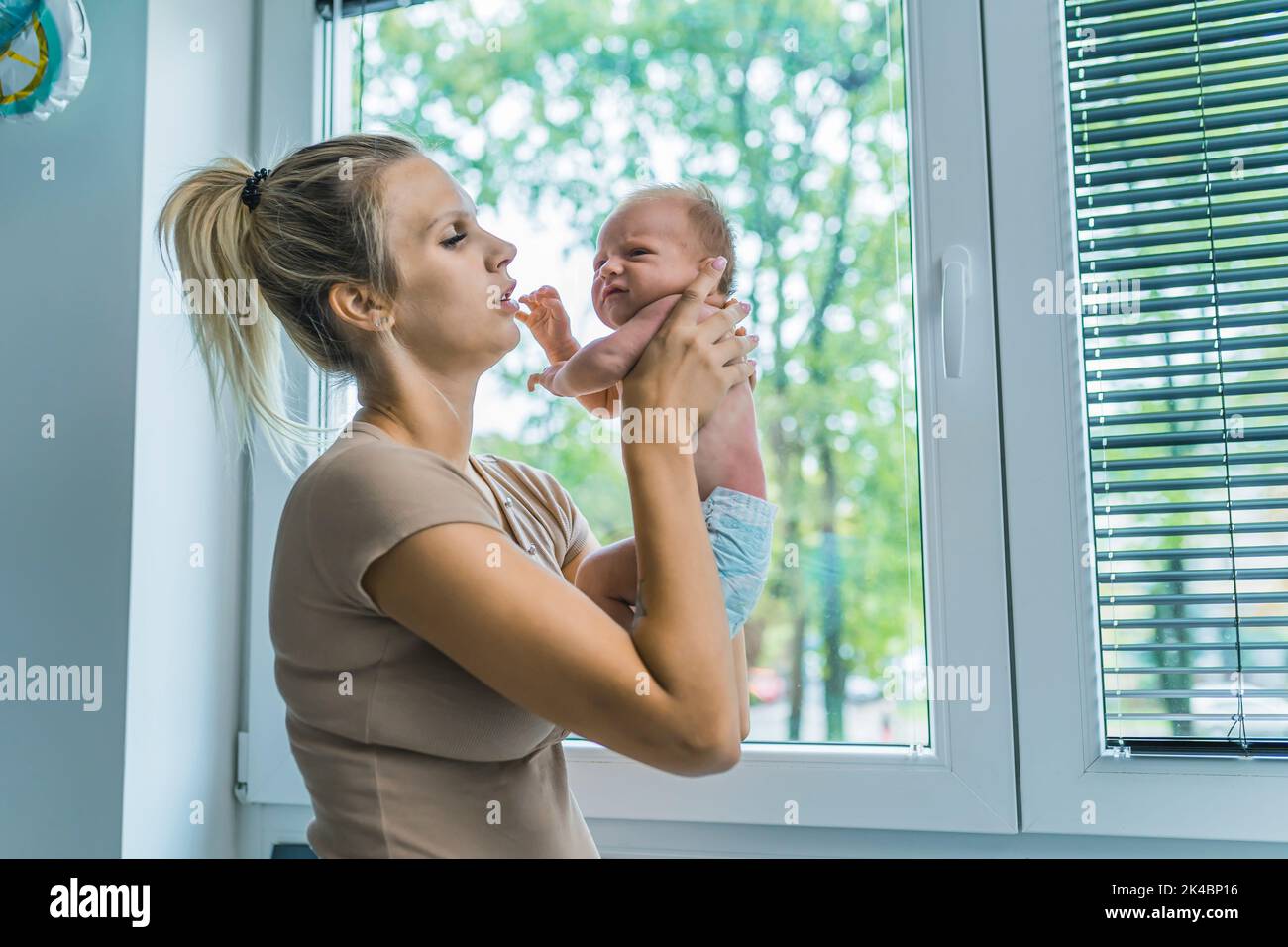 Caucasian happy loving mom holding her tiny newborn son in the air while standing near the window at home. High quality photo Stock Photo