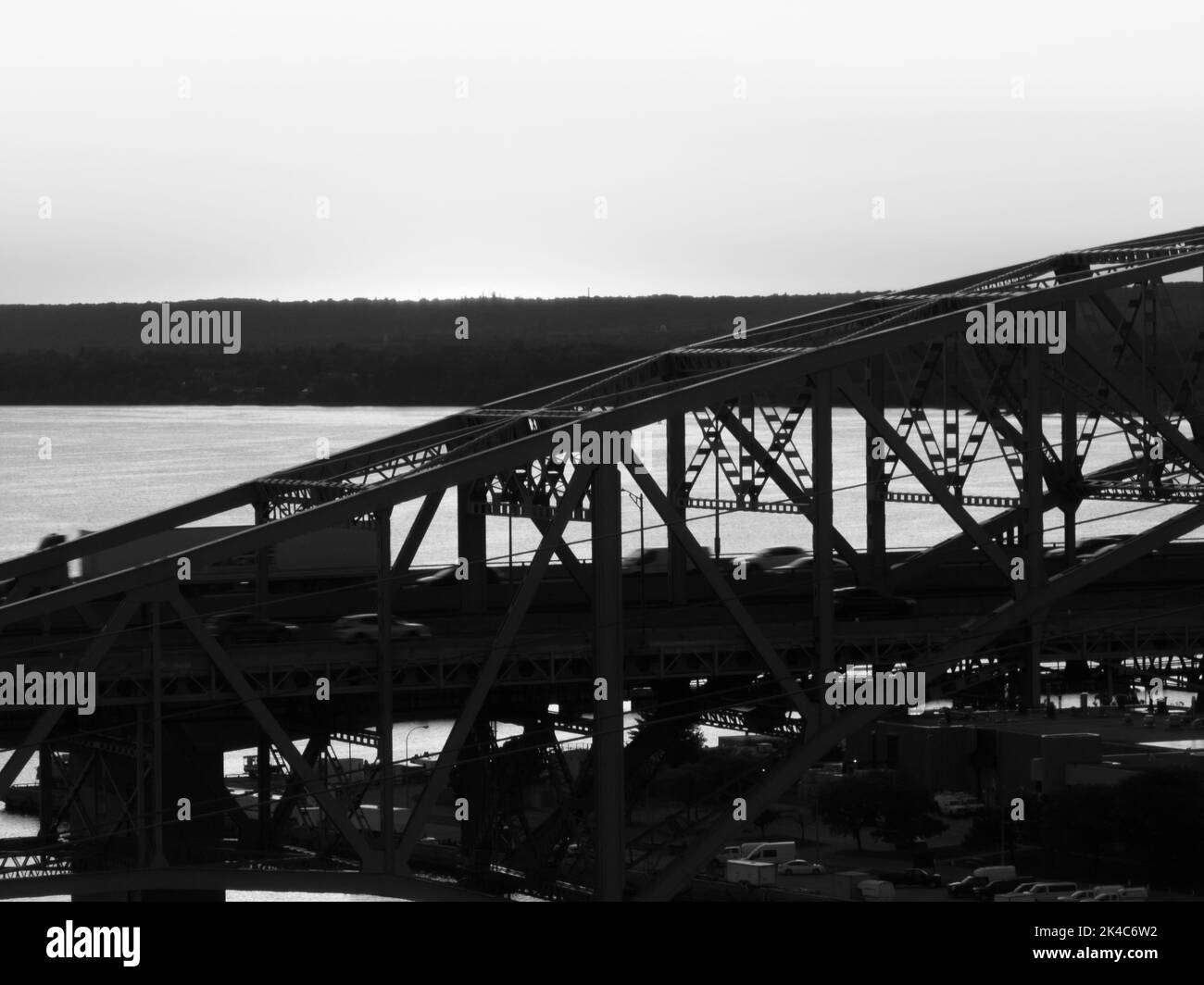 A black and white photo of a truss bridge over water, as traffic, motion blurred, travels across. Stock Photo