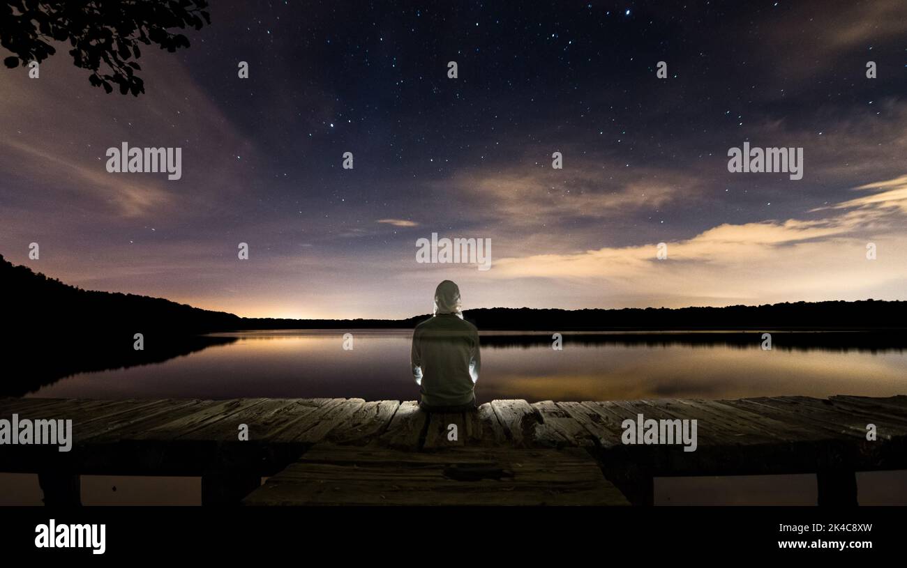 A beautiful shot of a person sitting on a wooden dock over a lake under a galaxy starry sky Stock Photo