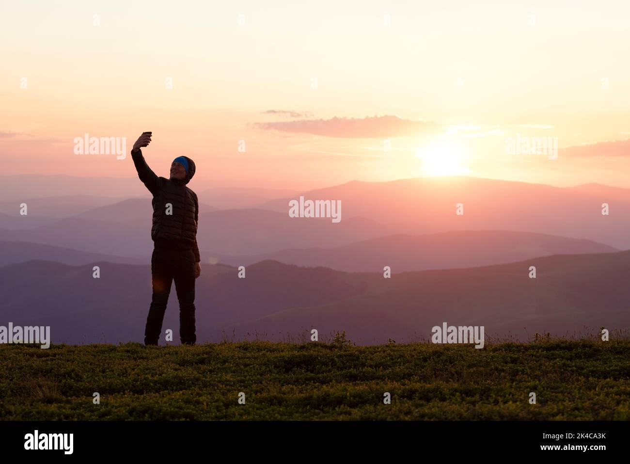 Tourist stands on top of a mountain taking a selfie in the colorful background at sunset Stock Photo
