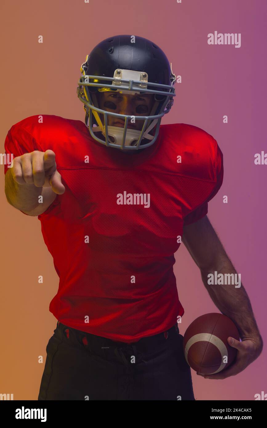 Caucasian male american football player holding ball with neon yellow and purple lighting. Sport, movement, training and active lifestyle concept. Stock Photo