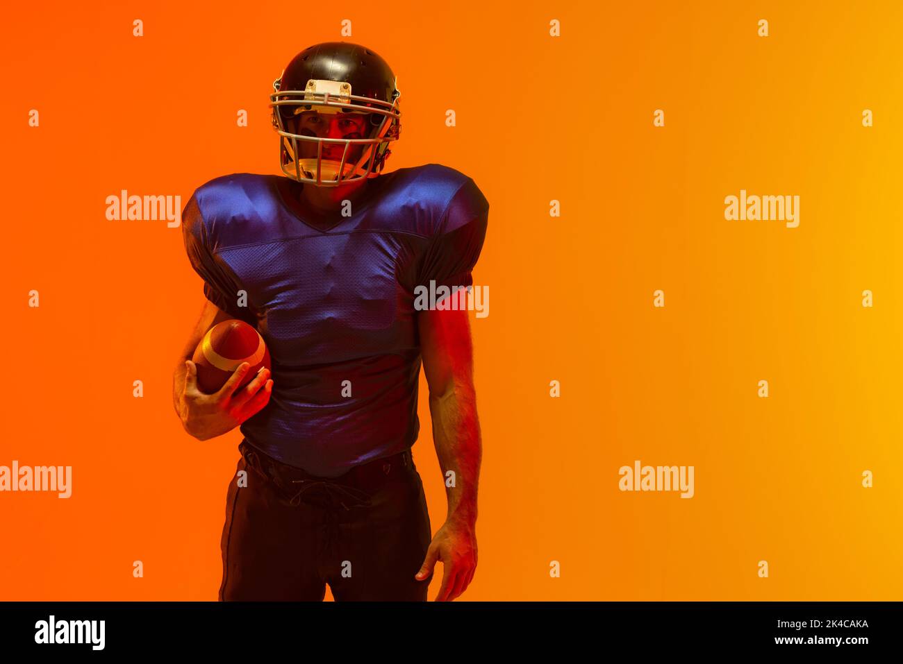 Caucasian male american football player holding ball with neon orange lighting. Sport, movement, training and active lifestyle concept. Stock Photo